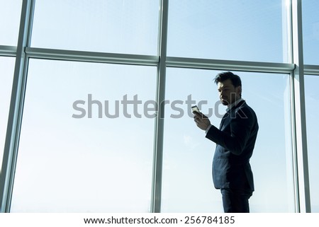 Side view of young businessman is standing near a panoramic window and looking at the phone.