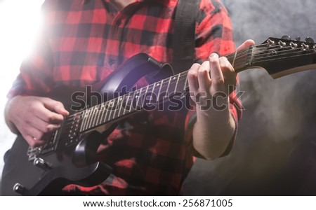 Close-up hands of young guitarist with the electric guitar, in dark room with light behind him.