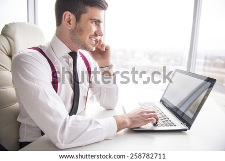 Smiling  businessman is working on his laptop in his office and is speaking phone and looking away.