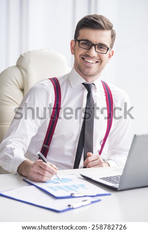 Handsome businessman is working with laptop and graphs in his office and looking at the camera.