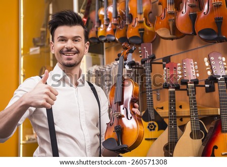 Young man is holding a violin and is showing thumbs up in a music store.