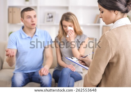 Psychologist talking with couple on the couch and taking notes in therapists office.