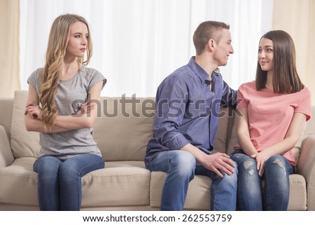 Jealousy. Young sad women sitting on the couch with her arms crossed while another women and men hugging near her.