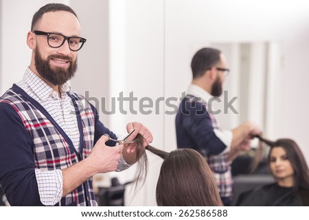 Male hairdresser making a haircut for a beautiful brunette girl in professional hairdressing salon and looking at camera.