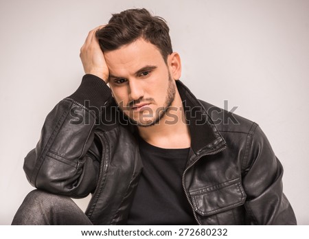 Young, serious man in leather jacket is sitting on the gray background and looking down.
