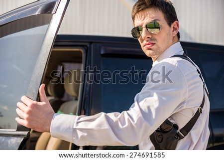 Young confident  businessman  in sunglasses with gun coming out of his luxurious car.