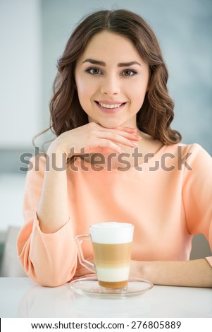 Portrait of smiling girl in peach blouse  sitting at the table with glass of coffee on grey background.