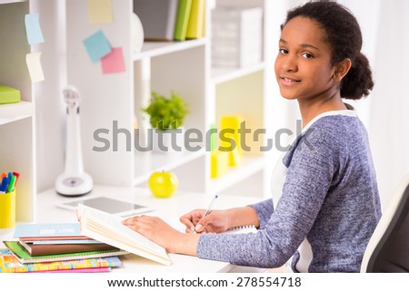 Young pretty schoolgirl sitting at the table and writing homework on colorful background.