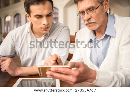 Two handsome businessmen in shirts sitting at the wooden table in urban cafe and using phone.