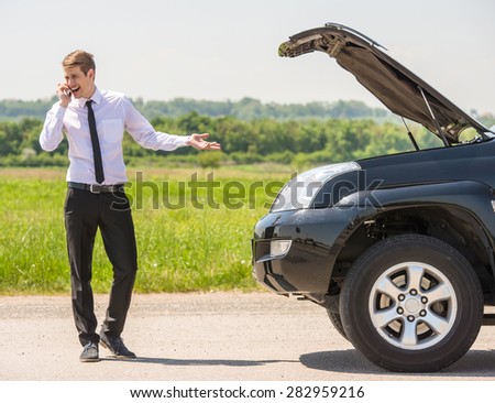 Young man having trouble with his broken car, opening hood and calling for help on cell phone.