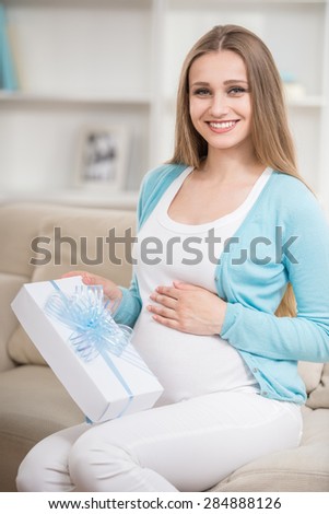 Beautiful pregnant woman sitting at sofa and holding gift.
