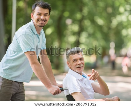Adult son walking with disabled father in wheelchair at park.