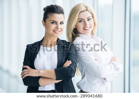 Two elegant female office workers standing back to back and smiling to camera.
