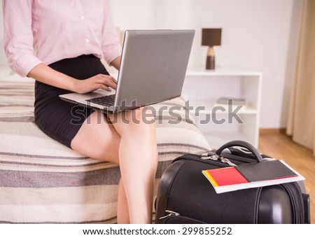 Business woman using her laptop while sitting on bed near suitcase and documents at the hotel room. Close-up.