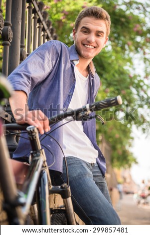 Young beautiful man standing with bicycle at the city street near the fence and looking at camera.
