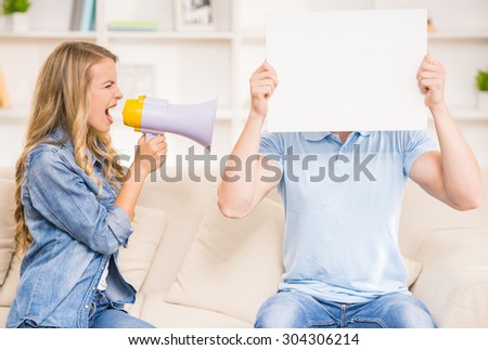 Wife shouting on her husband with mouthpiece while he closing his face with piece of paper.