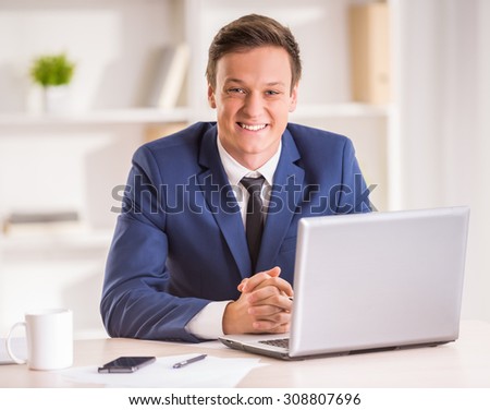 Young smiling businessman is looking at the camera while sitting with laptop in office