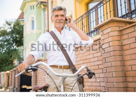 Senior man is talking by phone standing with bike in the street. Concept of active life elderly people.