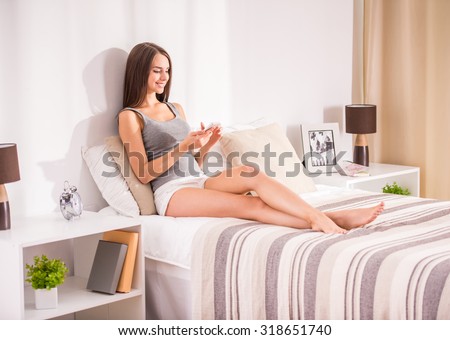 Young beautiful woman is sitting on the bed at home and looking at the smartphone.