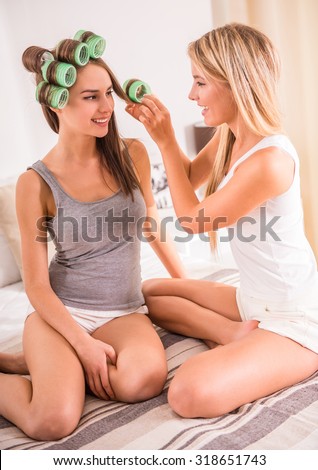 Two young female women with colorful hair rollers sitting on bed and smiling.