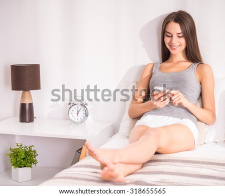 Young beautiful woman is sitting on the bed at home and looking at the smartphone.