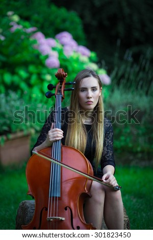 Pretty blonde girl in black dress playing a cello in a rural outdoor