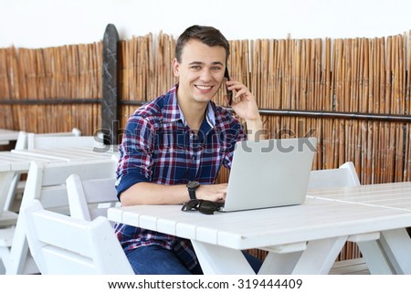Successful smiling freelancer talks on his smartphone while work on laptop computer in tropic cafe, stylish hipster use modern device