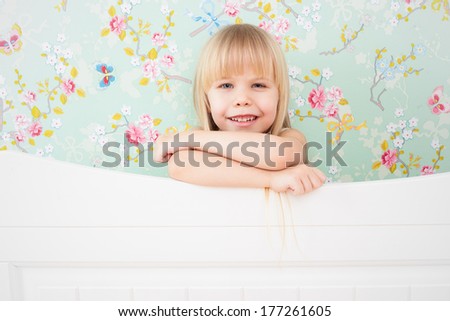 Little adorable girl with a sly smile standing behind a bed