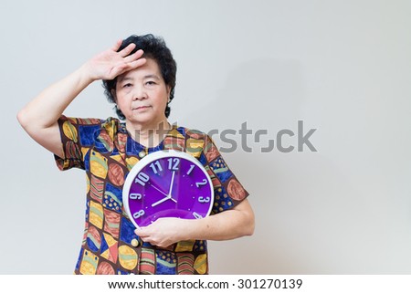 Asian senior woman showing and holding purple or violet clock in studio shot, on reddish yellow gray wall background with soft shadow