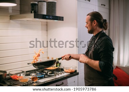 A male cook is cooking at the stove at home in the kitchen. A young guy in an apron in the kitchen at the gas stove fries food in a wok