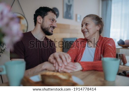The son congratulates his mother on the holiday, a family feast. An adult son cheerfully drinks tea with an elderly beautiful mother in the kitchen. A man visiting his beloved mother on a holiday
