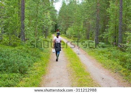A woman is walking with her dog in a forrest with a bucket of blue berries.