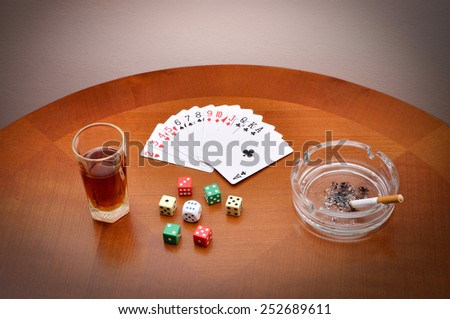 Glass of alcohol, cards, dice and ashtray with cigarette on the brown table