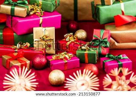 Many Christmas presents placed on a red cloth among baubles and stars. All gifts wrapped in magenta, gold, green and red. Each with unicolored ribbon. Focus on green bowknot around small red box.
