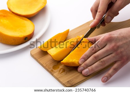 One third of a mango being subdivided into three chips. A left hand is keeping the fruit slice in place on a wooden board, while a right hand is cutting with a kitchen knife. White background.