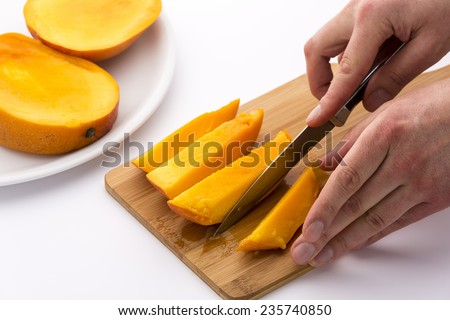 Pair of hands finished subdividing a mango third into four fruit chips. Knife and cutting board bedewed with the juice of this delicious tropical fruit. Two whole mango thirds waiting on white plate.