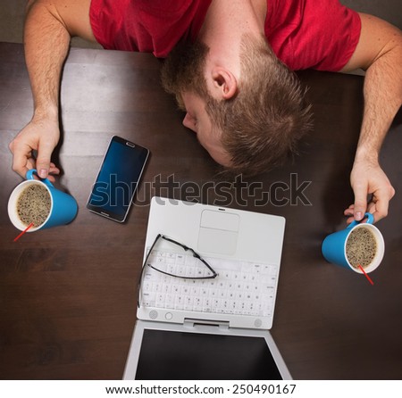 man sleeping on tabletop while holding cups of coffee while working at a desk on laptop