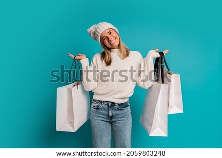 Studio winter photo of  smiling blond woman in white hat and whool sweater  a holding  shopping bags ,posing in studio over turquose background. 