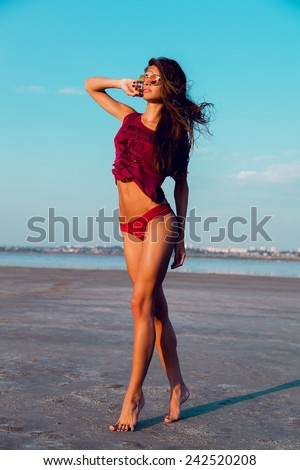 Young pretty fashion fitness lady posing outdoor in summer on the beach in hot weather in bikini and red t-short. Wearing stylish sunglasses .Summer mood.