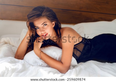Perfect sexy tan woman with  perfect skin , big full lips posing at bedroom, wearing sexy luxury stylish  black lingerie.l Lady lying on her stomach on white bed.