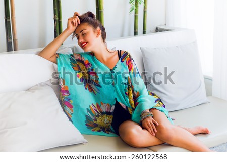 Elegant sexy woman  in colorful boho outfit  relaxing on white sofa in luxury hotel . Lady smile  and enjoying  holidays.