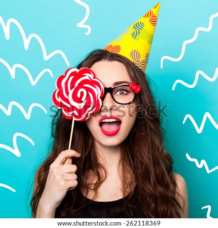 Studio closeup colorful portrait of young sexy funny fashion crazy woman posing on blue wall background in summer style outfit with pink lollipop wearing paper hat and cute glasses.