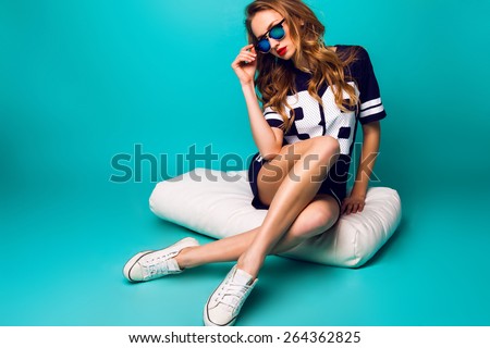 Close up fashion portrait of pretty sportive woman in stylish sunglasses, basketball t-short and white sneakers sitting on white pillow against bright aqua background .