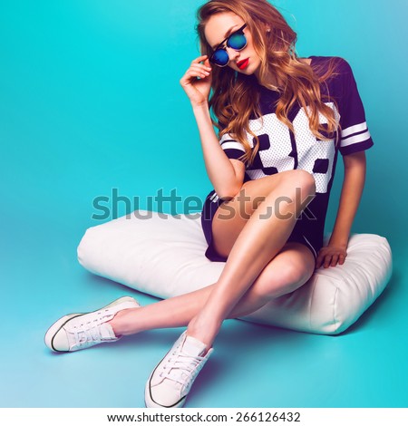 Close up fashion portrait of pretty sportive lady  in stylish sunglasses, basketball t-short and white sneakers sitting on white pillow against bright aqua background .
