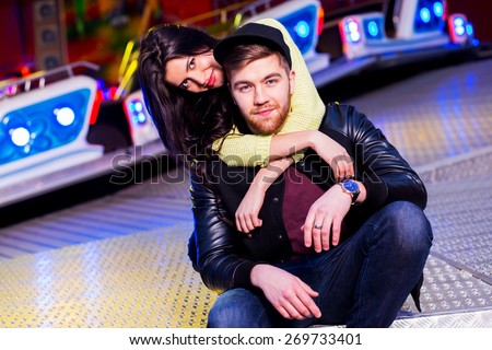 Joyful young stylish  couple being playful while visiting an attractions park arcade with rides. Have fun , enjoying  their time. Wearing spring trendy  outfit , neon sweater and swag hat.