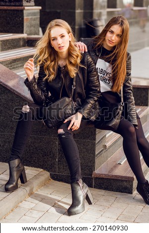 Two stylish  pretty  girls  posing on the street in sunny day. Wearing trendy urban outfit , leather jacket and boots heels.