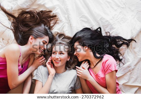 Top view of three best friends tell secrets in  bed . Group of pretty  young  women  in pajamas  lie on back and  talk.  Sunny warm colors. Cozy atmosphere.
