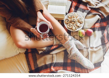Soft cozy photo of slim tan woman in warm sweater on the bed with cup of tea in hands, top view point. Girl sitting on checkered plaid near old books and cookies.