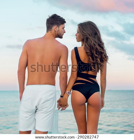 Young attractive  couple in love,  Handsome  man and sexy  woman with perfect tan slim fit  body  enjoying romantic evening on the beach, Holding hands watching  on the sea.