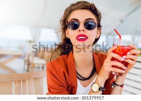 Close up lifestyle  portrait of cute pretty stylish young woman posing outdoor, sitting in  cafe and drinking  exotic cocktail, sea background. Bright colors. Vacation mood. Smiling and have fun.